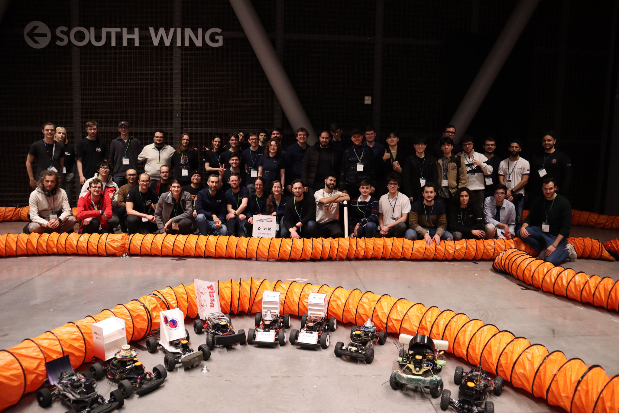 Roboracer participants gathered behind the orange track barriers, with autonomous cars in the foreground, at CDC 2024.