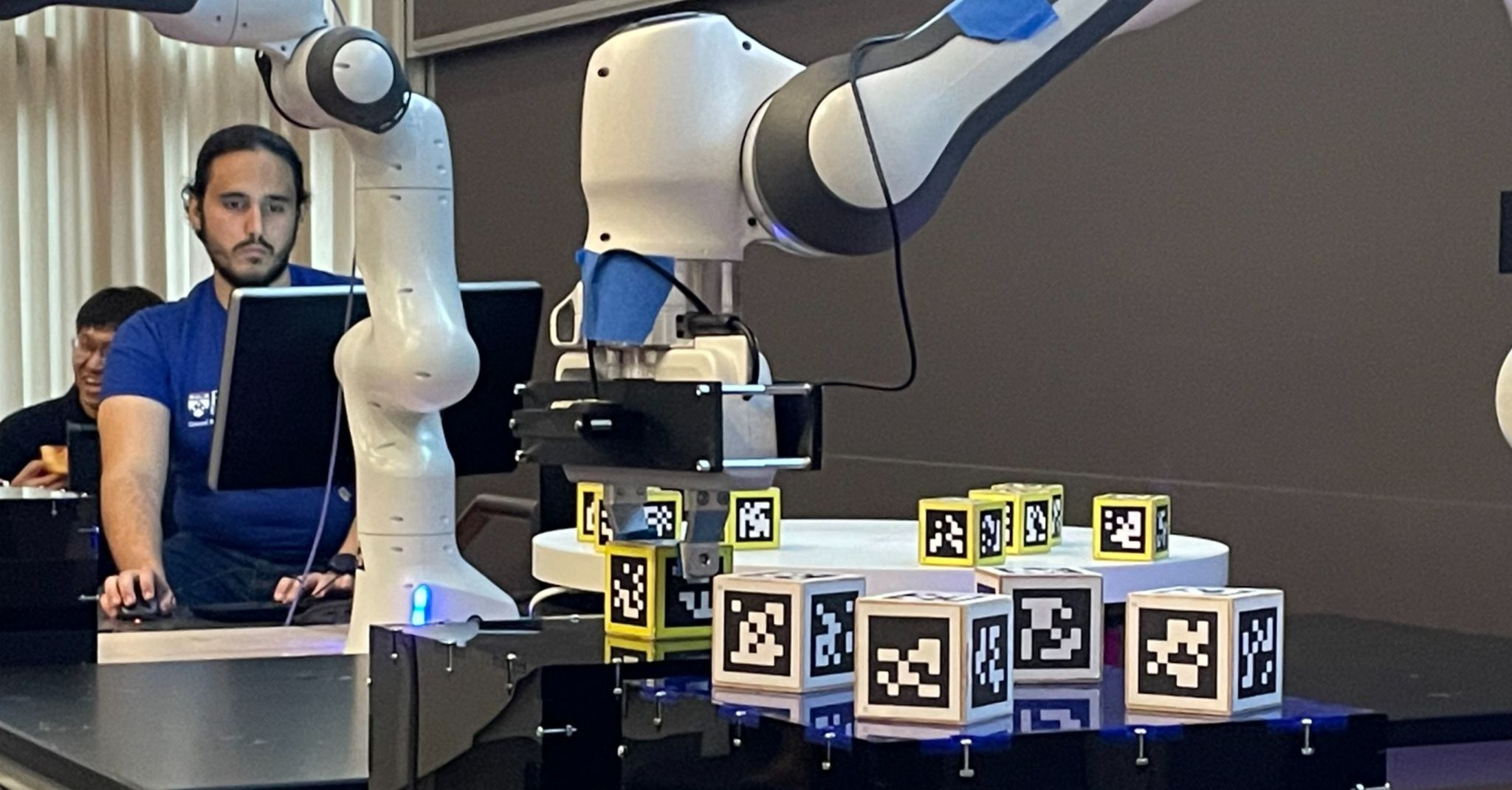 Ahmad working at a computer behind Franka Panda robot arms and blocks marked with AprilTags.